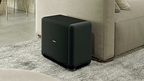 Modern black subwoofer next to a couch in a living room.