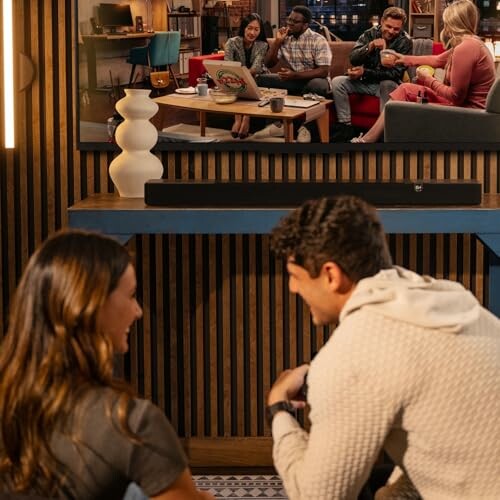 Two people sitting on the floor watching TV, with a group of friends on the screen.