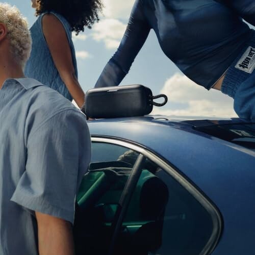 Three people in stylish outfits around a blue car with a small black speaker on top